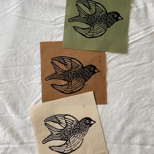 May include: Three square fabric patches with a black bird linocut print. The patches are in olive green, tan, and cream colors. The bird is depicted in flight with detailed wing feathers.