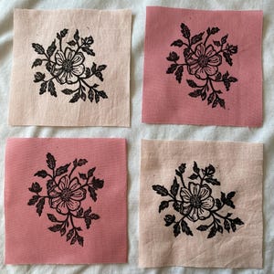May include: Four square fabric patches with a floral design. Each patch features a black floral print on a different colored background: two are pink, and two are a light peach color. The floral design includes a flower and leaves.