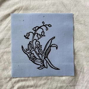 May include: A square, light blue fabric patch featuring a black linocut print of lily of the valley flowers. The design includes bell-shaped flowers, leaves, and stems, creating a botanical illustration.