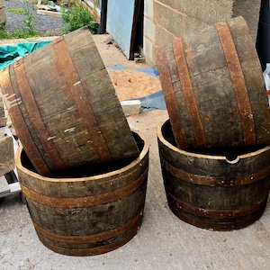 May include: Two weathered wooden barrels with metal bands. The barrels are dark brown with visible wood grain and rusty metal accents. One barrel has its top partially inside, while the other has its top fully inside. The barrels are sitting on a concrete surface.