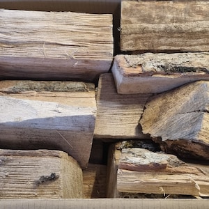 May include: A cardboard box containing split firewood. The logs are of various sizes and shapes, with visible bark and a natural, weathered appearance. The wood is light brown and tan in colour.