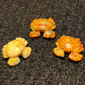 May include: Three small crab figurines. The crabs are carved from stone and feature a variety of colors, including yellow, orange, and brown. The crabs have detailed legs and claws. The figurines are displayed on a dark, textured surface.