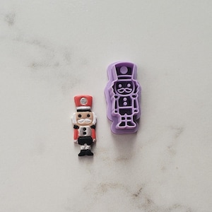 May include: A small nutcracker figurine with a red hat, white mustache, and black uniform, positioned next to a light purple plastic nutcracker-shaped cookie cutter. Both items feature the same soldier-like design with a tall hat and uniform.