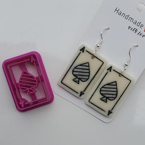 Puede incluir: Pendientes hechos a mano con el diseño de la carta del As de Picas, con ganchos plateados. También se ve un cortador de galletas rosa con la forma del As de Picas. Los pendientes son blancos con detalles negros. La tarjeta dice "Handmade with love".