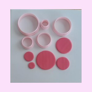May include: A collection of five light pink circular cookie cutters of varying sizes and four solid dark pink circular fondant or clay discs of corresponding sizes arranged in a group on a flat surface for baking or crafting projects.