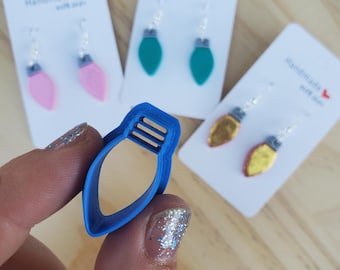 May include: A blue plastic light bulb-shaped cookie cutter is held in front of three pairs of handmade earrings. The earrings are shaped like Christmas lights in pink, teal, and gold. Each pair is attached to a white card.