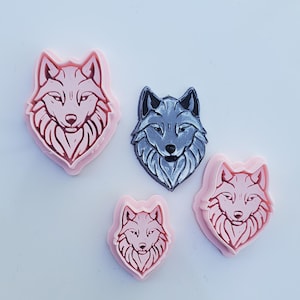 May include: Four wolf-shaped cookie cutters. Three are pink, and one is gray with black accents. The wolf heads have detailed facial features and are on a white surface.