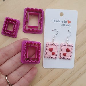 May include: Pink heart-shaped earrings and matching cookie cutters. The earrings have a pink glitter border and red hearts. The cookie cutters are square with a scalloped edge. The card reads "Handmade with love."