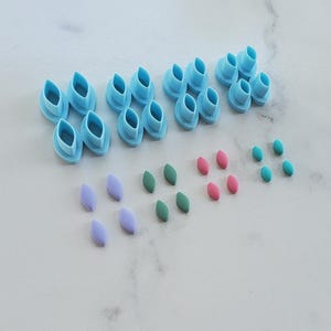 May include: A set of four light blue plastic petal-shaped cutters arranged in a row, accompanied by sixteen small, smooth, marquise-shaped pieces in lavender, dark green, pink, and teal, laid out in four distinct color-coordinated groups.