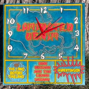 May include: A blue and yellow wooden clock with a red clock hand. The clock face features a blue and white map with the text "Laminated Denim". The clock is divided into three sections with the text "The Land Before Timeland", "King Gizzard and the Lizard Wizard", and "Hypertension".
