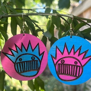 May include: A pair of wooden earrings with a blue and pink cartoon face design. The earrings are round and have a gold hook.