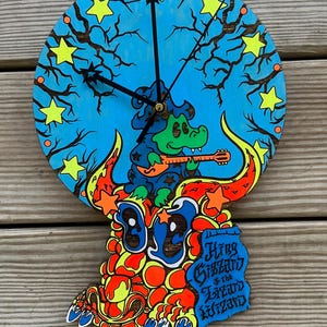 May include: A round, wooden clock with a vibrant, cartoonish design. The clock face is blue with a tree and star motif. A green alligator plays a guitar. The clock hands are black. The text "King Gizzard & the Lizard Wizard" is on the clock.