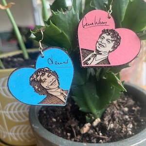 May include: A pair of heart-shaped wooden earrings with portraits of Gene Ween and Dean Ween. The earrings are painted in pink and blue.