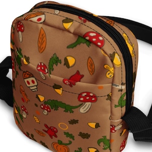 May include: A brown canvas crossbody bag with a black zipper and strap. The bag features a repeating pattern of mushrooms, acorns, leaves, and other woodland creatures.