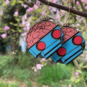 Puede incluir: Un par de pendientes de madera con forma de cerebro con un diseño azul y rosa. Los pendientes están colgados de una rama con flores rosas.