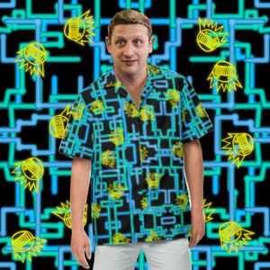 May include: A man wearing a black, blue, and yellow short-sleeved shirt with a geometric pattern. The shirt has a button-down collar and a relaxed fit.