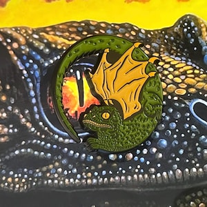 Puede incluir: Pin de esmalte verde y amarillo de un dragón con alas enroscadas alrededor de su cuerpo. El dragón tiene un diseño texturizado y escamoso.