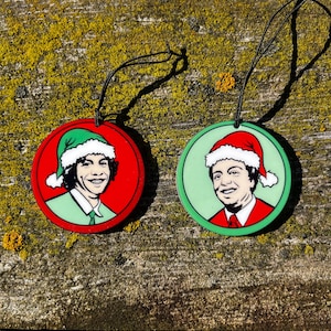 Puede incluir: Dos adornos navideños redondos con retratos de dibujos animados de una persona con gorro de Papá Noel. Uno tiene un fondo rojo con un borde verde, el otro tiene un fondo verde con un borde rojo. Ambos tienen cuerdas negras para colgar.