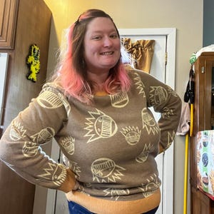 May include: A brown sweater with a repeating cream-colored graphic pattern. The sweater has a crew neck and ribbed cuffs and hem in a contrasting orange color. The person in the image is wearing blue jeans.