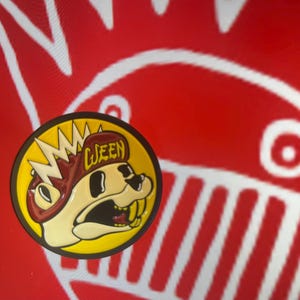 May include: A yellow and red enamel pin featuring a cartoon character with a red hat that says "Queen" and a mohawk. The character has a white face and a black mouth with teeth.