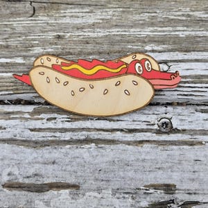 Puede incluir: Un recorte de madera con temática de hot dog con un diseño de dibujos animados. El pan del hot dog es de color marrón claro con semillas de sésamo, y el hot dog tiene detalles rojos y amarillos. El hot dog tiene una cabeza de cocodrilo.