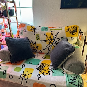 May include: A white armchair with a colorful throw blanket featuring a retro geometric pattern. The blanket has a repeating design of stars, squares, and circles in shades of yellow, orange, green, and blue. Two gray throw pillows are on the chair.