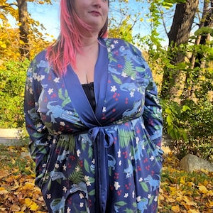 May include: A navy blue robe with a whimsical alligator print in shades of blue and green. The robe features a tie waist, long sleeves, and pockets. White floral and red berry accents complete the design. The robe is worn outdoors.