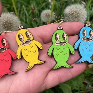 Puede incluir: Cuatro pendientes de madera en forma de pez de color rojo, amarillo, verde y azul. Cada pez tiene una cara de dibujos animados con ojos grandes y una boca pequeña.