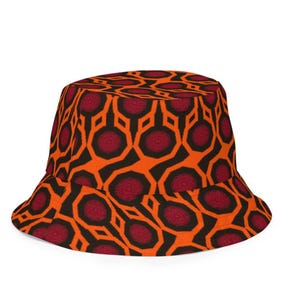 May include: Bucket hat with a repeating geometric pattern in orange, black, and burgundy. The hat features a wide brim and a rounded crown. The pattern consists of interconnected shapes with circular accents.