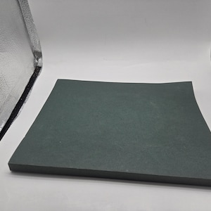 May include: A dark gray, square-shaped cutting board. The board is made of a solid material and has a slightly curved edge. The board is set against a white background.