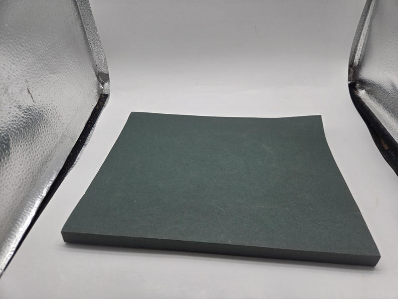 May include: A dark gray, square-shaped cutting board. The board is made of a solid material and has a slightly curved edge. The board is set against a white background.