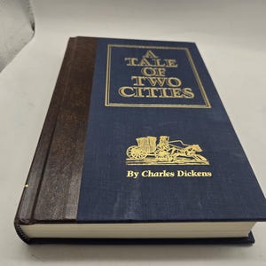 May include: Hardcover book titled "A Tale of Two Cities" by Charles Dickens. The book has a navy blue cover with gold lettering and a gold illustration of a horse-drawn carriage. The spine is brown.