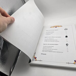 May include: A book with white pages, open to reveal the table of contents. The book has decorative corner designs and a detailed list of chapters. A hand is turning a page, showing the book's interior.