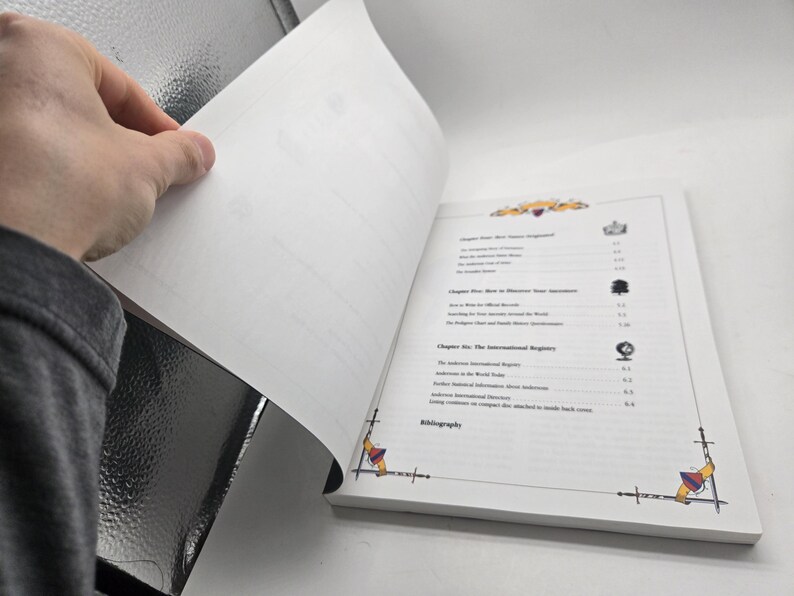 May include: A book with white pages, open to reveal the table of contents. The book has decorative corner designs and a detailed list of chapters. A hand is turning a page, showing the book's interior.
