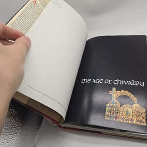 May include: An open book with the title "the age of chivalry" on a black page. The book features a gold and jewel-encrusted crown illustration. The opposite page is blank. The book's pages are white with text.