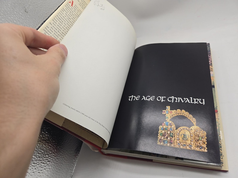 May include: An open book with the title "the age of chivalry" on a black page. The book features a gold and jewel-encrusted crown illustration. The opposite page is blank. The book's pages are white with text.