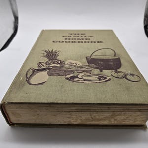 May include: A vintage cookbook titled "The Family Home Cookbook" with a green cover. The cover features illustrations of a pineapple, vegetables, a pitcher, and a pot. The book is slightly worn, suggesting it's an older edition.