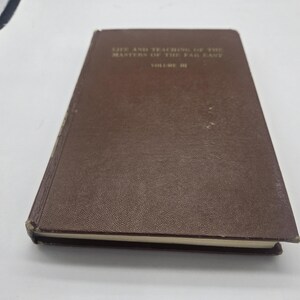 Peut inclure: Un livre à couverture rigide marron intitulé "Life and Teaching of the Masters of the Far East, Volume III". Le livre a une couverture texturée et semble être vintage. Le titre est imprimé en lettres dorées.