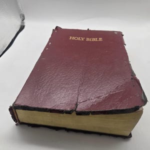 Könnte beinhalten: Eine kastanienbraune Heilige Bibel mit strukturiertem Einband und goldfarbenem Schriftzug auf der Vorderseite. Das Buch weist sichtbare Abnutzungserscheinungen an den Kanten auf, mit ausgefranstem Material und freiliegender Bindung, sowie Goldschnitt.