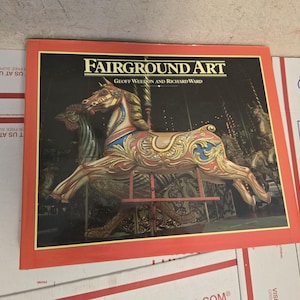 Könnte beinhalten: Ein Buch mit dem Titel "Fairground Art" mit rotem Rand. Das Cover zeigt ein detailliertes Bild eines Karussellpferdes mit goldenen, blauen und roten Akzenten. Die Namen der Autoren, Geoff Weeron und Richard Ward, sind ebenfalls sichtbar.