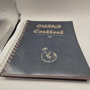Puede incluir: Un libro de cocina encuadernado en espiral titulado "OHANA Cookbook" con el subtítulo "Recetas hawaianas culturalmente diversas". La portada es azul oscuro con texto blanco y un gráfico de una isla con una palmera.