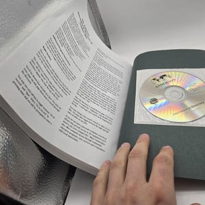 May include: An open book with printed text and a CD-ROM. The CD-ROM is in a clear plastic sleeve and has the text "ANDERSONS" and "International Directory" printed on it. The book's pages are white, and the cover is black.