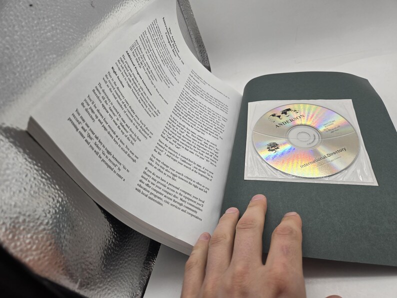 May include: An open book with printed text and a CD-ROM. The CD-ROM is in a clear plastic sleeve and has the text "ANDERSONS" and "International Directory" printed on it. The book's pages are white, and the cover is black.