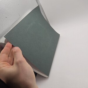 May include: A hand lifting a gray sheet of paper from a stack of similar sheets. The paper has a slightly textured surface and a muted, neutral color. The background is a mix of white and a reflective, textured surface.