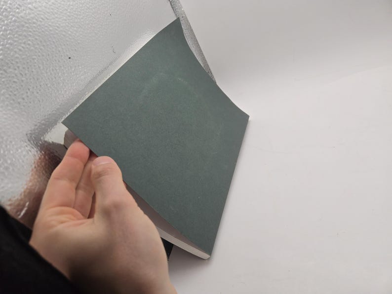 May include: A hand lifting a gray sheet of paper from a stack of similar sheets. The paper has a slightly textured surface and a muted, neutral color. The background is a mix of white and a reflective, textured surface.