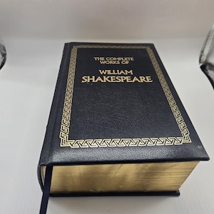 May include: A hardcover book titled "The Complete Works of William Shakespeare." The book has a black cover with gold lettering and a decorative gold border. The edges of the pages are gold. A blue ribbon bookmark is visible.