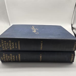 Könnte beinhalten: Zwei marineblaue Hardcover-Bücher mit dem Titel "My Experiences in the World War" von Pershing. Band I und Band II sind sichtbar, mit goldener Schrift und einer Unterschrift auf dem oberen Buch. Der Verlag ist Stokes.