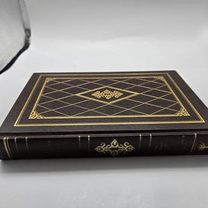 May include: A dark brown hardcover book featuring a gold foil diamond grid pattern on the front cover with a central decorative filigree element. The spine includes gold horizontal lines and a matching central filigree design with text.