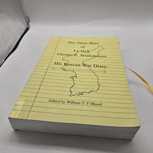 Puede incluir: Un libro de tapa blanda con cubierta de color amarillo pálido y líneas horizontales negras que recuerdan al papel de cuaderno. El título es The Three Wars of Lt. Gen. George E. Stratemeyer His Korean War Diary, con un mapa de Corea.