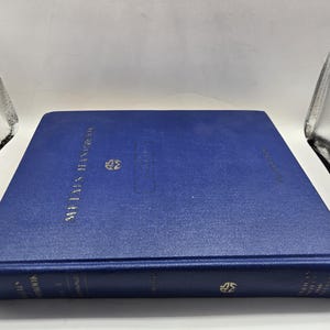 Puede incluir: Un libro de tapa dura azul con letras doradas y un emblema dorado. El libro se titula "Metals Handbook" y parece ser una guía de referencia. El libro está tumbado sobre una superficie blanca.